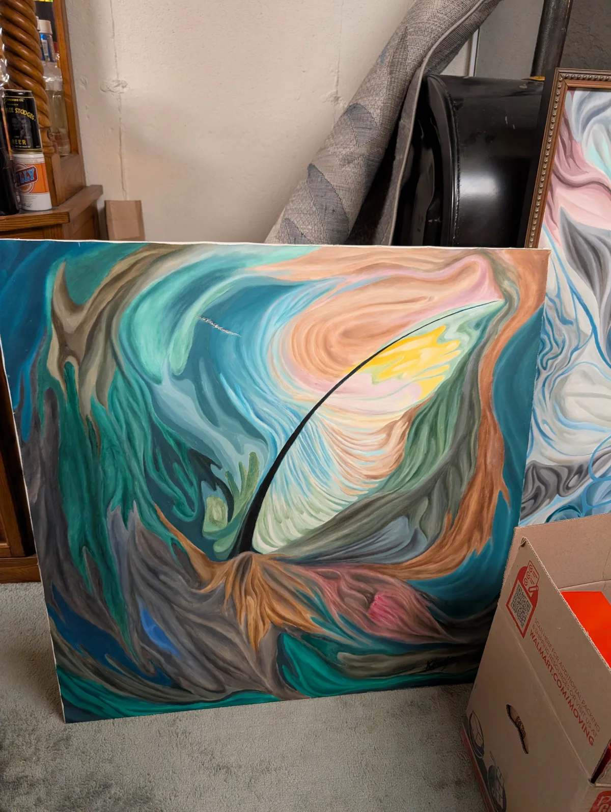 John King's *Middle East* (1989), an oil on canvas featuring swirling, biomorphic forms in deep teals, warm ochres, pinks, and soft greens that spiral around a luminous yellow-white center, bisected by a sweeping black arc. The fluid, organic composition evokes turbulent natural forces—wind, water, or geological upheaval—rendered with sinuous, interlocking passages of color. A second, similarly styled painting is partially visible at right. The unframed canvas is propped against furniture in a storage setting.